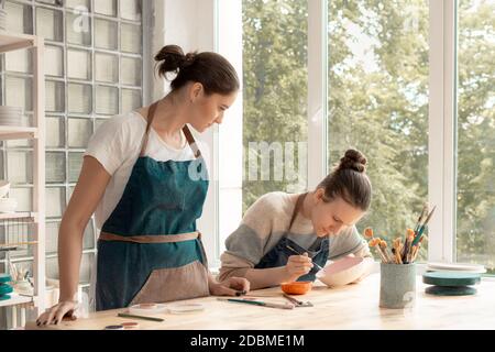 Donna che fa ceramica. Attraente giovane donna esperta in grembiule in piedi al tavolo e l'insegnamento Foto Stock