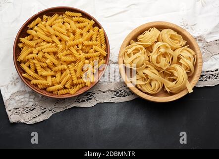 fusilli crudi e fettuccine in piatti di legno su un tavolo nero, vista dall'alto Foto Stock