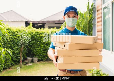 Asian giovane consegna uomo corriere invio e tenuta fast food pizza inscatolato in uniforme lui protettivo viso cliente servizio a casa porta, sotto il cur Foto Stock