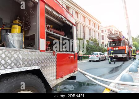 Vigili del fuoco che indossano uniforme in piedi dietro nastro antincendio e molti incendi carrelli motore con scaletta in caso di incidente nella torre alta residenziale o edificio di uffici Foto Stock