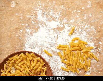 pasta di fusilli grezza in un piatto marrone di ceramica su un tavolo di legno con farina bianca spruzzata, vista dall'alto Foto Stock