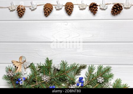Garland con coni e angeli, rami di pino con ornamenti in legno e bacche blu su sfondo di legno bianco. Modello scheda anno nuovo, spazio per la copia Foto Stock