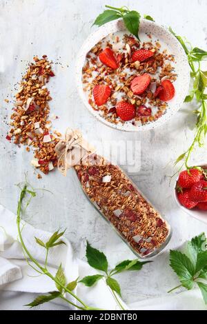 Granola in una bottiglia di vetro su uno sfondo chiaro. Fragole con latte e muesli su un piatto. I grani sono sparsi sulla tabella. Il concetto di a. Foto Stock