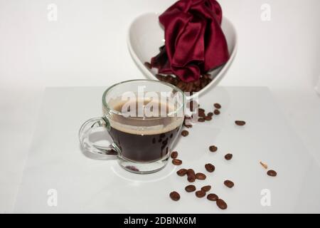 Tazza di vetro con caffè nero con schiuma galleggiante il liquido accanto ad un fazzoletto rosso e il caffè torrefatto fagioli contenuti in una ceramica bianca he Foto Stock