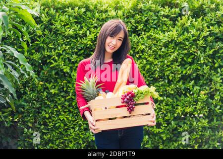 Ritratto di Asian bella giovane donna contadina in piedi sorride e tenendo pieno cibo fresco frutta verdura cruda in una scatola di legno nelle sue mani sul verde Foto Stock