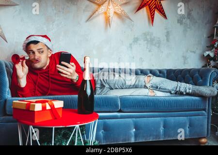 Giovane bell'uomo in cappello Babbo Natale con bicchiere di champagne che prende selfie sullo smartphone. Ragazzo divertente che si fotografa sul telefono cellulare e mentendo o Foto Stock