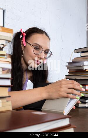 Una giovane ragazza si siede educato in una scrivania, tenendo una lettura della scuola nelle sue mani e davanti a lei ci sono due mucchi di libri da leggere. Foto Stock