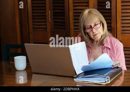 Donna matura che lavora con un computer portatile e carte da guardare. È indoor in un tavolo di legno con un computer argento. Tazza di caffè da parte. Foto Stock