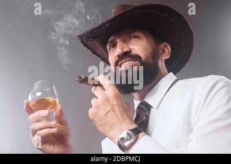 Il cowboy con la bearded sicuro sta fumando il sigaro e bevendo l'alcool su fondo grigio. Foto Stock