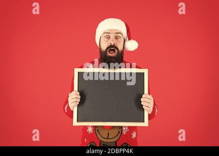 Incredibile. Annuncio d'inverno. Evento invernale. Vacanze invernali. Man tenere lavagna vuota copia spazio. Guy babbo natale sfondo rosso. Santa Claus hat con la pubblicità. Lavagna per informazioni. Foto Stock