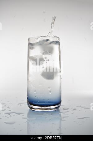 Un cubo di ghiaccio che cade in un bicchiere d'acqua creando uno spruzzi. Il bicchiere ha un colore blu alla base con spruzzi d'acqua che circondano la bevanda. Foto Stock