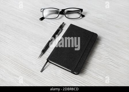 Foto di blocco note nero chiuso, bicchieri e matita meccanica su sfondo tavolo in legno chiaro. Modello per posizionare il progetto. Foto Stock