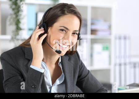 Donna di telemarketer felice con la cuffia che si pone guardando la fotocamera seduto su una scrivania in ufficio Foto Stock