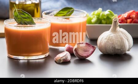 Gazpacho in due bicchieri, con i suoi ingredienti, pomodoro, aglio, olio, pepe rosso e pepe verde. Foto Stock