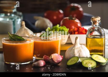 Gazpacho in due bicchieri, con i suoi ingredienti, pomodoro, aglio, olio, cetriolo, pepe rosso e pepe verde. Foto Stock