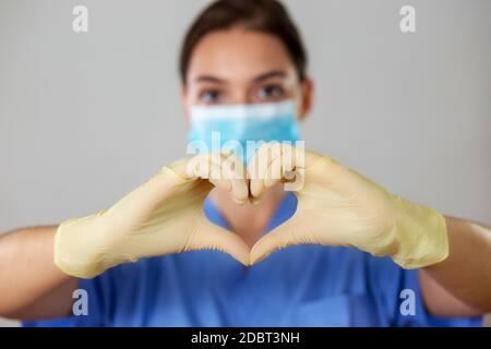 Giovane ragazza in una maschera medica che mostra il simbolo di un cuore con le mani guanto davanti a lei su uno sfondo di studio luminoso. Vista frontale del caucasico Foto Stock