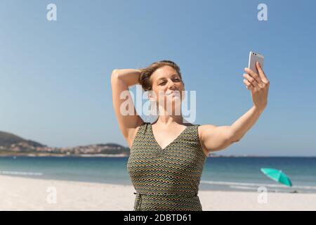 Donna bionda di mezza età in un vestito verde che fa un selfie sulla spiaggia con il mare sullo sfondo Foto Stock