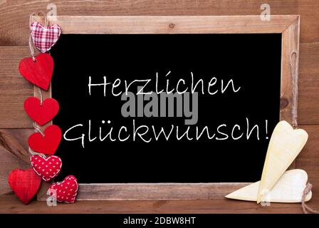 Balckboard con testo tedesco Herzlichen Glueckwunsch significa Complimenti. Decorazione del cuore rosso e giallo. Marrone legno sfondo Foto Stock