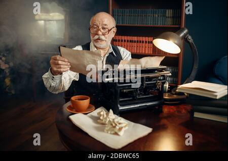 Anziano scrittore lavora su macchina da scrivere vintage nel suo ufficio domestico. L'uomo anziano in occhiali scrive romanzo di letteratura in camera con fumo Foto Stock