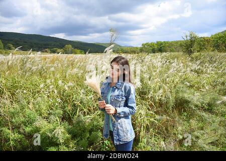 Giovane donna sorridente che tiene in mano un bouquet di Miscanthus sinensis (erba d'argento cinese) e lo guarda sullo sfondo di un campo. Au Foto Stock