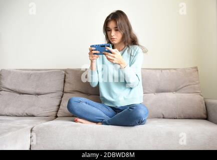 Sorpresa giovane donna che tiene smartphone con due mani, seduta sul divano in abiti casual. Sta leggendo nuovi giochi o giocando. Attività di blocco Foto Stock