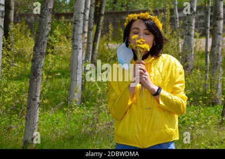 Donna bruna con occhi chiusi, in giacca gialla con corona di dandelioni e rimossa maschera medica odore bouquet di dandelioni. pande coronavirus Foto Stock