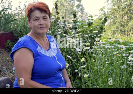 Ritratto di donna anziana in abito blu seduta accanto a. aiuole di fiori con margherite in villaggio in una serata estiva Foto Stock