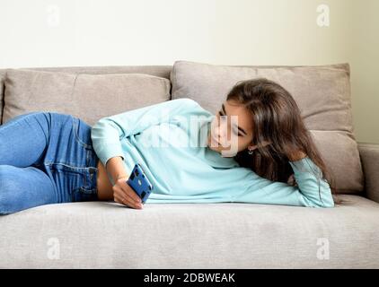 Annoiata giovane donna sdraiata sul divano in abiti casual, appoggiando la mano sotto la testa. Tiene e scorre lo smartphone Foto Stock