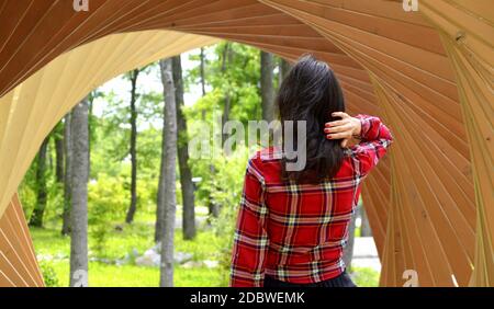 Donna bruna in camicia rossa in piedi con la schiena alla macchina fotografica e tenendo i capelli in mano all'interno del tunnel di legno con archi. Percorso alla foresta Foto Stock