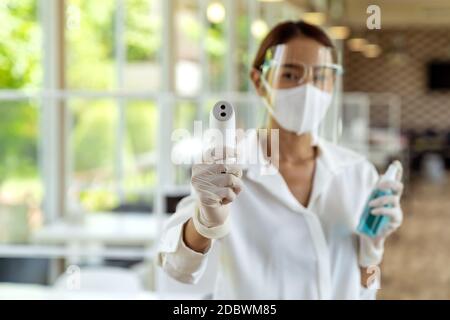 Ritratto attraente cameriera asiatica indossare maschera viso e visiera tenere termometro e igienizzatore in gel di alcol per la pulizia e l'igiene con riposo interno Foto Stock