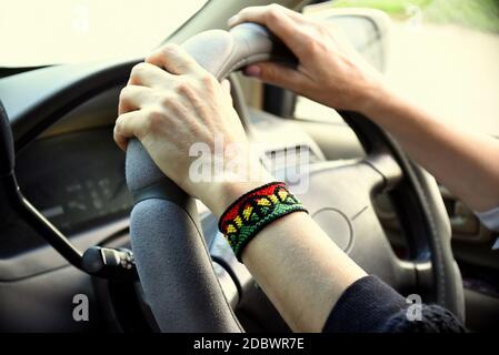 Braccialetto fai da te con motivo Rasta flag e simbolo di pace sul polso femminile. Mani che tengono il volante nella vettura. Foto Stock