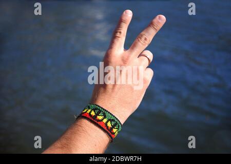 Fuoco selettivo del bracciale di amicizia fai da te fatto a mano con motivo bandiera Rasta con un simbolo di pace sul polso maschile su sfondo mare. Foto Stock