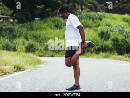 Primo piano Asian giovane atleta sportivo corridore uomo nero indossare orologio piedi sollevamento gambe e ginocchio, prima di correre al parco di salute strada all'aperto, lui Foto Stock