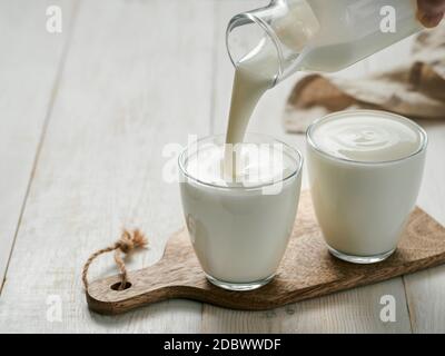 Versando kefir, latticello o yogurt fatti in casa con probiotici. Yogurt che scorre da una bottiglia di vetro su sfondo bianco di legno. Probiotico freddo fermentato da Foto Stock