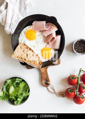 Uova fritte in padella nera con verdure, pane e prosciutto su sfondo chiaro. Vista dall'alto Foto Stock