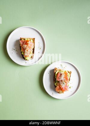 Sanwiches sani aperti con pane di segale scuro, avocado, salmone affumicato e germogli su piatti su sfondo verde, vista dall'alto, spazio di copia Foto Stock