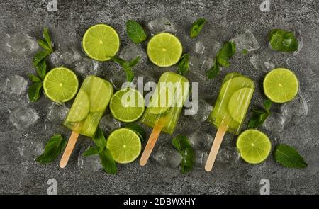 Primo piano di diversi papsicli surgelati di succo di frutta con fette di lime fresche, foglie di menta verde e cubetti di ghiaccio sulla superficie grigia del tavolo, vista dall'alto, diretta Foto Stock