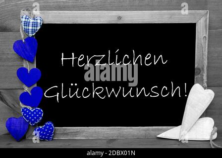 Balckboard con testo tedesco Herzlichen Glueckwunsch significa Complimenti. Sfondo grigio in legno con decorazione colorata cuore blu Foto Stock