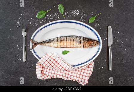 Pesce di sgombro affumicato intero su piatto ovale bianco, pietra nera grigia come tabella sotto, sale di roccia e insalata di foglie verdi vicino. Vista dall'alto. Foto Stock