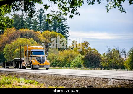 Carro lungo giallo brillante Big RIG rosso industriale semi-camion trasporto di un semirimorchio a gradino vuoto in marcia rettilinea strada autostradale con verde Foto Stock