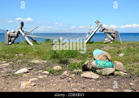 Iscrizione Aloha dipinta su un mucchio di pietre su uno sfondo di amaca fatta in casa con bastoni di legno e pietre. Mare giapponese in una giornata estiva soleggiata. Foto Stock