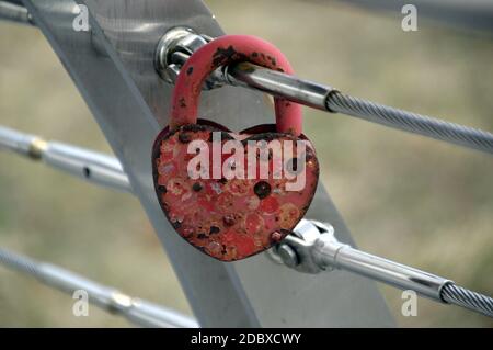 Vecchio lucchetto rosso arrugginito a forma di cuore appeso su una recinzione in metallo all'aperto Foto Stock