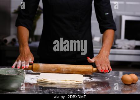 Lo chef prepara dolci in una cucina professionale. Impastare l'impasto. Uova, farina, tondino. Sfondo scuro. Foto Stock