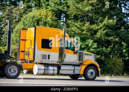 Carro lungo giallo brillante Big RIG rosso industriale semi-camion trasporto di un semirimorchio a gradino vuoto in marcia rettilinea strada autostradale con verde Foto Stock