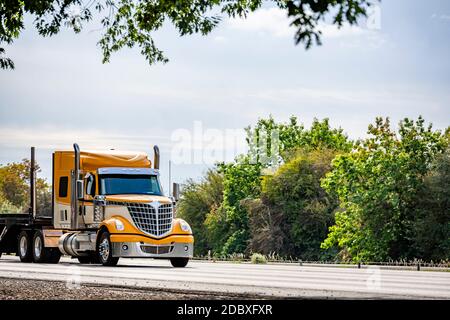 Carro lungo giallo brillante Big RIG rosso industriale semi-camion trasporto di un semirimorchio a gradino vuoto in marcia rettilinea strada autostradale con verde Foto Stock