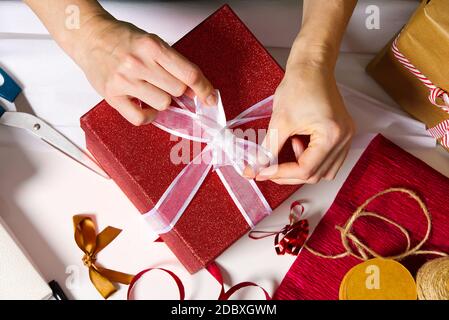 le mani della donna imballano un regalo di natale in una scatola rossa con un nastro bianco. imballaggio del regalo di diy Foto Stock