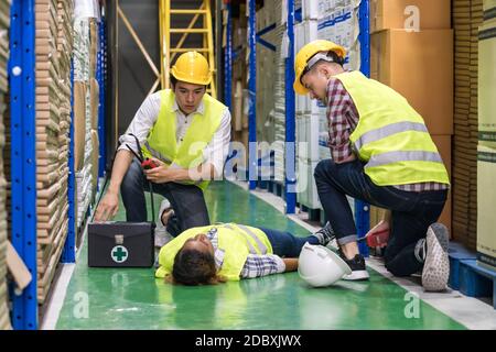 Il lavoratore del magazzino si è subito assistito al collega che si trova sul pavimento del magazzino dopo un incidente durante il lavoro. Utilizzo per la sicurezza industriale First e busin Foto Stock