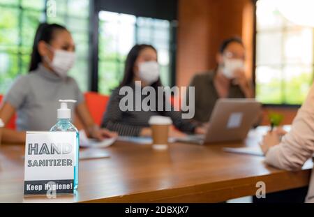 Segnaletica di gel alcolico igienizzante per le mani nella sala riunioni con sfondo di team asiatici che lavorano e si scambiano idee. Indossano una maschera facciale nuova Foto Stock