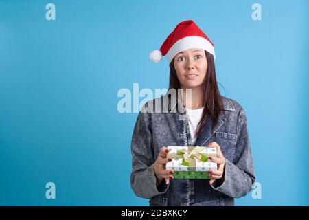 Primo piano di una giovane donna asiatica disgustata e sconvolta in un cappello di Babbo Natale che tiene una scatola regalo verde, su uno sfondo blu Foto Stock