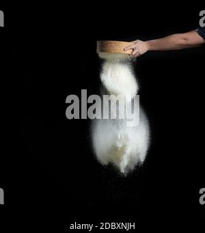 femmina mano setaccia farina di grano bianco attraverso un setaccio di legno rotondo su uno sfondo nero, il prodotto viene versato Foto Stock